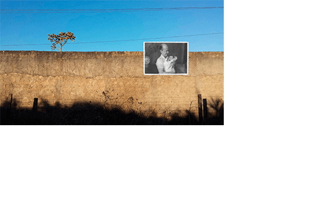 Foto horizontal de um muro de cimento visto de frente, iluminado pelo sol, com uma sombra bem escura ao longo de toda a parte de baixo. Acima, um céu azul sem nuvens e alguns fios da rede elétrica. Por trás do muro, à esquerda, vê-se o topo de uma árvore pequenininha, com as folhas secas. À direita há uma foto antiga em preto e branco, pequena, colada sobre o muro e o céu. Ela mostra um velho careca de camisa clara, sorrindo de leve e segurando um bebê novinho, enrolado numa manta branca.