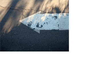 Foto horizontal de uma seta branca pintada no asfalto, apontando para a esquerda. Na parte de cima da foto há sol, e a de baixo está na sombra. Sombras de capim se alongam em direção ao alto da imagem. Na parte ensolarada há uma rachadura horizontal que atravessa toda a foto, descendo ligeiramente e passando por cima da seta, que está quase toda na sombra. 