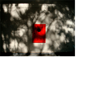 Foto horizontal de uma parede com um retângulo pintado de vermelho vivo, visto de frente, no centro da imagem. Ele é vertical e tem um buraco redondo na parte de cima. A parede é bege claro. Toda a imagem está coberta por sombras que fazem uma espécie de rendilhado quase preto na parede ensolarada; parecem sombras de árvores. Em cima há uma estreita faixa preta onde o sol não bate, e embaixo uma estreita faixa cinza escura de piso.