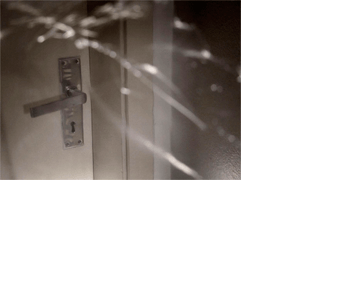 Foto horizontal da maçaneta barata de uma porta comum, branca, fechada, e um pedaço da parede. A foto é bastante escura. O anteparo na lente faz vários traços desfocados e brancos sobre quase toda a imagem. Os traços lembram rachaduras.