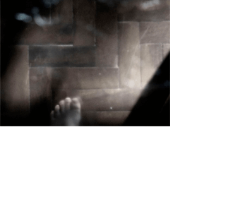Foto horizontal de metade de um pé esquerdo descalço sobre um chão de tacos. O pé está embaixo, no centro, um pouco à esquerda. No canto direito há uma sombra formando um triângulo preto até quase em cima. A imagem é escura e está toda desfocada pelo anteparo na lente.