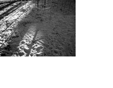 O chão de areia branca de parque infantil ocupa todo o quadro, com uma faixa de luz do sol na metade da esquerda. No alto, à direita, vê-se a parte de baixo de um trepa-trepa, que lança sombras diagonais sobre a parte iluminada.