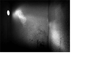 Foto de um aposento vazio, muito pequeno e escuro, muito apertado. Vê-se a parede do fundo e as laterais. Na parede da esquerda, a mais estreita e mais escura, há uma pequena abertura redonda por onde entra um pouco de luz. As paredes estão manchadas e inteiramente cobertas por pequenos buracos feitos manualmente.
