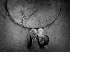 Detalhe da tampa de um bueiro e de duas botas velhas sobre um chão de cimento. O bueiro forma um semicírculo na parte de cima da foto, e está tampado. Na parte de baixo, ao centro, há duas botas de couro escuro. A ponta das botas toca a tampa do bueiro. Elas estão muito usadas e arranhadas, sem cadarços e manchadas com um pó branco.