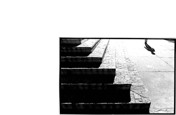 Foto horizontal mostrando os pés de um homem que anda em direção à câmera, no canto de cima, à direita. A sombra de todo o seu corpo se alonga à sua frente. Há uma escadaria em L fazendo uma linha diagonal que atravessa a foto de alto a baixo, da esquerda para a direita. Acima dessa diagonal o chão está claro, quase branco; abaixo, está quase preto por causa da sombra dos degraus. Na imagem vê-se apenas seis degraus da escada, a larga calçada de cimento e a sombra do homem.