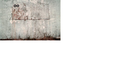 Foto horizontal; detalhe uma parede branca desgastada pelo tempo, que agora está cinza e escura, com muitas manchas. No centro da parede havia uma janela larga que foi toda tapada com tijolos, exceto por dois cobogós pequenos, no canto superior esquerdo. Essa janela tapada foi parcialmente rebocada e pintada de branço; vê-se vestígios dos tijolos vermelhos por baixo. No chão há uma faixa estreita de mato seco.