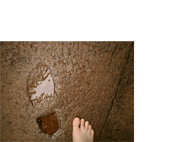 Foto horizontal mostrando o detalhe de uma calçada de cimento com uma pegada antiga, feita por um sapato ou bota quando o cimento estava fresco. A calçada está molhada e a pegada está cheia de água. Ela aparede do meio para a esquerda da foto, e à sua direita há metade de um pé descalço.