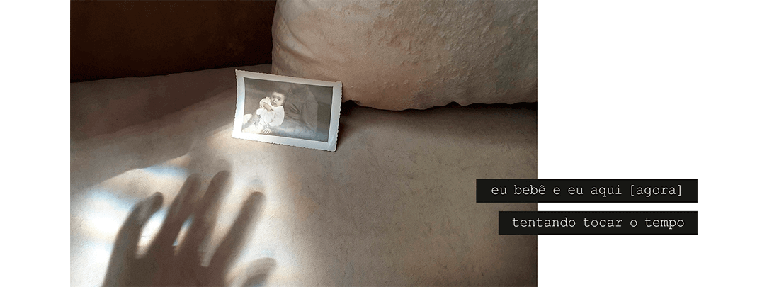 Foto horizontal e escura, em tons de bege e marrom, mostrando a sombra de uma mão. Ela vai em direção a uma pequena foto antiga em preto e branco, encostada na almofada de um sofá. Essa foto mostra um bebê, que parece olhar espantado e interessado para a câmera. Na foto maior, uma faixa de luz do sol em formato triangular sai da esquerda e vai até a foto menor, com a ponta chegando no bebê. A sombra da mão está sobre essa parte iluminada. Há uma frase escrita em duas faixas pretas que começam à direita e ultrapassam a margem da foto: eu bebê e eu aqui [ agora ] tentando tocar o tempo.