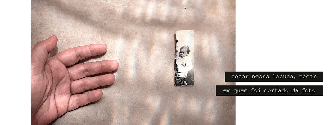 Foto horizontal com fundo bege claro, algumas sombras e desenhos da luz que vem de cima. No lado direito há um pedaço de uma foto antiga, estreito, vertical e rasgado do lado. No canto esquerdo da foto maior aparecem os dedos e a palma de uma mão que parece tentar alcançar a foto menor. O pedaço de foto mostra um bebê mas não permite identificar a pessoa que o está segurando, pois foi rasgada. Há uma frase em duas faixas pretas que começam no lado direito e saem da margem: tocar nessa lacuna, tocar / em quem foi cortado da foto.