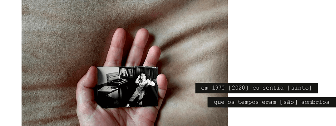 Foto horizontal mostrando uma mão virada para cima, segurando na palma uma foto pequena. A mão está no centro da imagem e pressiona levemente um fundo bege e macio, fazendo algumas rugas e sombras. A foto é antiga, em preto e branco, e mostra uma menina de três anos apoiada numa cadeira. Ela está em meio perfil e olha para a esquerda, séria. O lugar está bastante escuro; há uma estante com livros e uma mesinha com uma pequena vitrola. Uma frase sobre duas faixas pretas começa na parte direita e sai da margem: em 1970 [2020] eu sentia [sinto] que os tempos eram [são] sombrios.