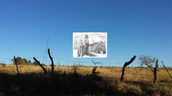 Foto horizontal mostrando em sua maior parte um céu muito azul, sem nuvens. Na parte de baixo há uma faixa de pasto com capim seco, iluminada pelo sol. Abaixo do pasto, uma faixa quase preta onde o sol não bate. No limite entre as duas há uma cerca de arame farpado frouxo; pode-se ver quatro estacas tortas, feitas com galhos de árvore. Estão escuras e suas pontas se destacam contra o céu azul. Bem no centro da foto, sobre o céu, está colada outra foto pequena, antiga, em preto e branco, mostrando uma menina de sete ou oito anos andando num velho cavalo branco, sem sela.