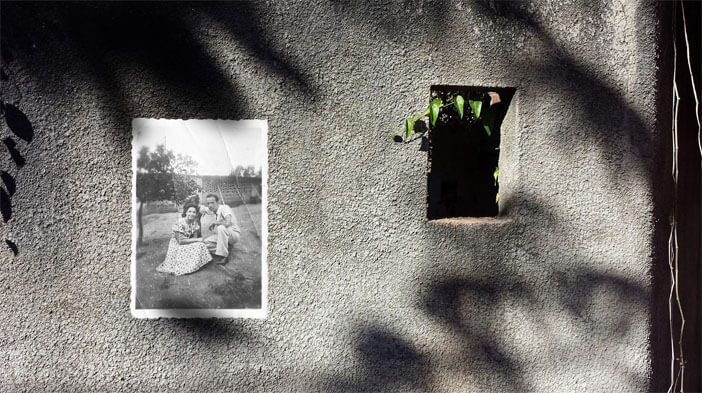 Foto horizontal de um muro de cimento visto de frente, iluminado pelo sol, com uma sombra bem escura ao longo de toda a parte de baixo. Acima, um céu azul sem nuvens e alguns fios da rede elétrica. Por trás do muro, à esquerda, vê-se o topo de uma árvore pequenininha, com as folhas secas. À direita há uma foto antiga em preto e branco, pequena, colada sobre o muro e o céu. Ela mostra um velho careca de camisa clara, sorrindo de leve e segurando um bebê novinho, enrolado numa manta branca.