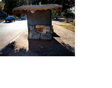 Foto horizontal de uma barraca de rua feita de pedaços de madeirite, com pintura azul velha e desbotada. A barraca está de frente; na parte de baixo lê-se uma palavra grande, que pode ter sido "sapateiro", mas está quase toda coberta por outras coisas escritas: dois números de celular, os nomes Jair e Vânia e alguns papéis ilegíveis. O teto é um velho toldo de lona, com beiral bem comprido, debaixo de uma árvore. A barraca está na calçada na beira de uma rua onde há dois carros passando. O sol vem de trás, de modo que a barraca lança uma grande sombra para a frente, que ocupa toda a parte de baixo da foto.