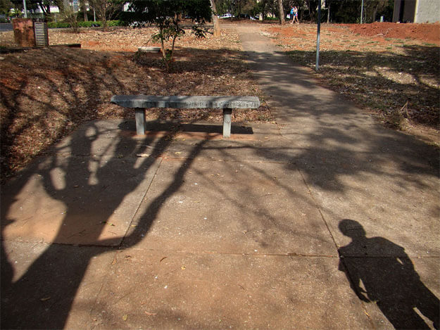 Foto horizontal que mostra a sombra de uma pessoa em pé, na diagonal, no canto da direita. Do canto esquerdo parte a sombra de uma árvore alta e sem folhas. Há um espaço cimentado em formato quadrado na maior parte da foto; ele continua numa calçada que vai subindo até o topo. Vê-se um banco de cimento sem encosto e, bem ao longe, duas pessoas passando. Há sombras de outras árvores e o chão está forrado de folhas secas. Tudo está em tons de marrom, bege e alguns pontos de terra vermelha.