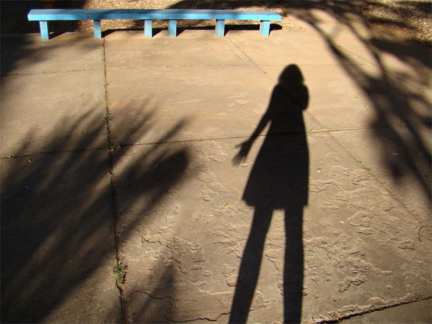 Foto horizontal da sombra de uma mulher de vestido, com o braço esquerdo um pouco afastado do corpo e a mão aberta. Em volta dela há outras sombras, margeando e entrando na foto. O chão é de cimento marrom. Bem no alto da foto vê-se um comprido banco de cimento azul turquesa, vivamente iluminado pelo sol. Atrás dele e no canto direito há um chão coberto de pequenas folhas secas.