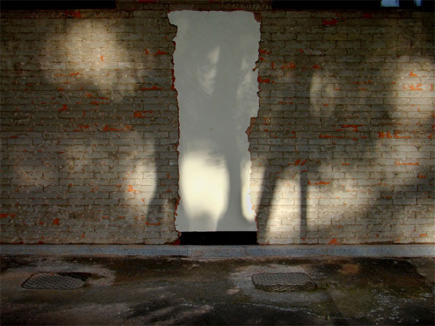 Foto horizontal de uma fachada de parede que foi toda descascada, mostrando velhos tijolos marrons e vermelhos. Bem no centro há uma área que não foi descascada, onde antes havia uma porta. A porta foi tampada com tijolo, rebocada e pintada de bege claro. O contorno dela é bem irregular. Há pequenas manchas de sol nessa não-porta e na parede em volta, mas na foto predominam as sombras. No chão de cimento em frente à não-porta há dois bueiros com tampa de ferro.