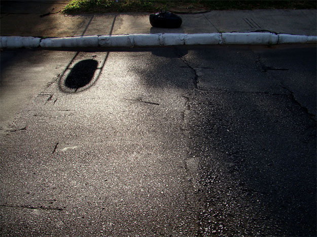 Foto horizontal mostrando um chão de asfalto em quase toda a imagem. Bem no alto há um meio fio branco velho e uma calçada, que atravessam a foto horizontalmente. Uma faixa de sol vem de cima, lançando no asfalto a sombra de um saco de lixo preto e uma sombra misteriosa de algo que não aparece na foto, uma estrutura comprida que sustenta uma forma arredondada.