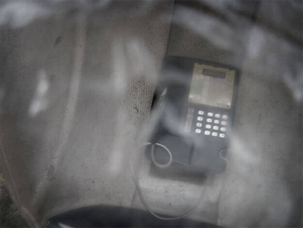 Foto horizontal mostrando a parte de dentro de um orelhão de rua, escuro, velho e sujo, com teclas prateadas claras que se destacam. A concha do orelhão é branca, também velha e suja. Toda a foto está desfocada pelo anteparo, menos uma área bem pequena do centro, entre a borda do aparelho e o fundo. O enquadramento pende ligeiramente para a direita.