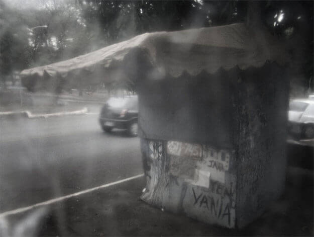 Foto horizontal de uma barraca de rua feita de pedaços de madeirite. Ela está de lado; na parte de baixo lê-se os nomes Jair e Vânia, em meio a papéis ilegíveis e manchas de tinta. O teto é um velho toldo de lona, com beiral bem comprido, e parede estar debaixo de uma árvore. A barraca está na calçada na beira de uma rua onde há um carro passando. Toda a imagem está desfocada pelo anteparo.