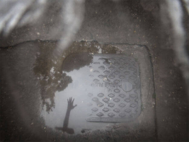 Foto horizontal de um bueiro quadrado com tampa de ferro sobre uma calçada de cimento. Está centralizado do meio para baixo. Há uma poça d'água sobre metade da tampa e parte da calçada. Nessa poça aparece, na parte de baixo, o reflexo escuro de um braço estendido, com a mão aberta, e da copa de uma árvore. A imagem é bem escura, exceto pela água. Está toda desfocada pelo anteparo, com algumas manchas brancas na parte de cima.