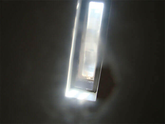 Foto horizontal de uma janela basculante com formato incomum, vertical, muito estreita e comprida. Está enquadrada de frente, no centro da foto, cortada pela margem de cima e se estendendo até quase embaixo. O enquadramento é torto e a janela se inclina para a direita. Ela deixa entrar a luz de fora. Em volta da janela há apenas uma parede no escuro, quase preta. Tudo está desfocado pelo anteparo, menos uma área que mostra a borda da janela.