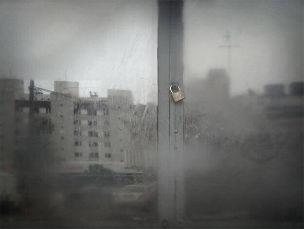 Foto horizontal com o detalhe de uma porta de vidro velha e suja, que reflete alguns prédios ao fundo. Há uma faixa de ferro cinza no centro, onde a porta se fecha; essa faixa atravessa a imagem na vertical, e no meio dela há um cadeado fechado. Toda a imagem está desfocada pelo anteparo na lente, exceto por uma pequena área onde se vê o cadeado.