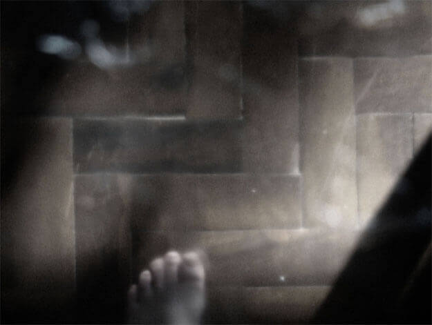 Foto horizontal de metade de um pé esquerdo descalço sobre um chão de tacos. O pé está embaixo, no centro, um pouco à esquerda. No canto direito há uma sombra formando um triângulo preto até quase em cima. A imagem é escura e está toda desfocada pelo anteparo na lente.