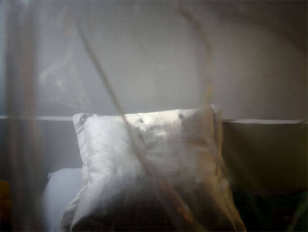 Foto horizontal de uma almofada branca apoiada na cabeceira reta e branca, contra uma parede branca. Ela é quadrada e está centralizada, colada à margem de baixo. O topo da almofada está iluminado pelo sol e o resto da imagem é escura. Ela está toda desfocada pelo anteparo na lente, que também faz duas linhas diagonais e escuras, de cima a baixo da foto, passando sobre a almofada.