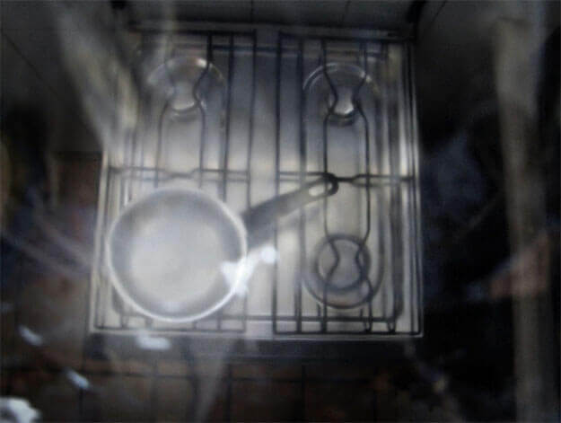 Foto horizontal de uma caçarola de alumínio sem tampa, que está sobre um fogão de quatro bocas com superfície prateada e trempe preta. A foto foi tirada do alto e o fogão faz uma forma quadrada um pouco torta no centro da imagem. Em volta dele tudo está escuro, quase preto. Toda a foto está desfocada pelo anteparo na lente, com manchas claras.