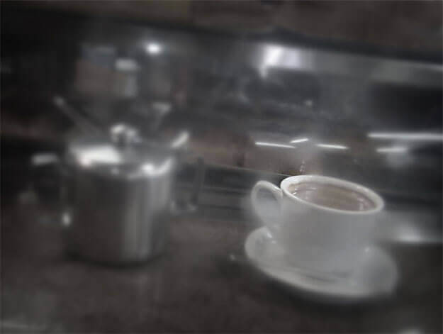 Foto horizontal de uma xícara branca e um açucareiro prateado sobre um balcão, vistos bem de perto. A imagem é bem escura e a xícara se destaca. Tudo está desfocado pelo anteparo na lente, menos uma área bem pequena da borda da xícara. O enquadramento faz o balcão pender ligeiramente para a direita.