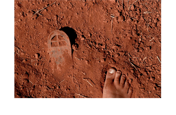 Série de 14 fotografias coloridas. Esta foto horizontal mostra o detalhe de um chão de terra vermelha e seca, com alguns torrões e pedacinhos de capim seco. No lado direito, em baixo, aparece metade de um pé descalço e muito sujo de terra. No lado esquerdo aparece a sola de um sapato, parciamente coberta de terra.