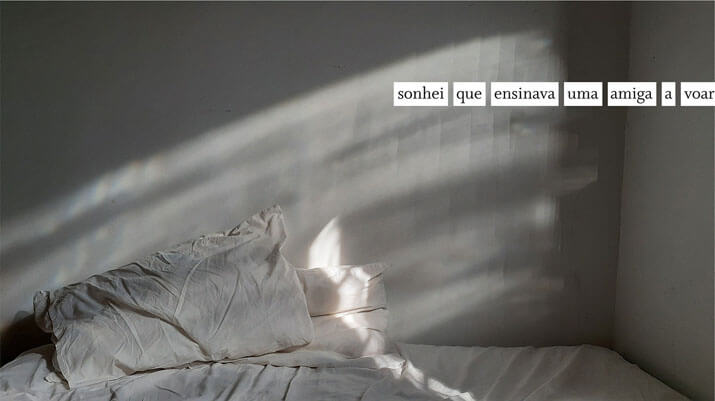 Foto horizontal mostrando duas paredes, um colchão e dois travesseiros, com fronhas e lençol brancos. A foto é escura e colorida mas parece em preto e branco, pois não há objetos coloridos. Uma luz filtrada pelo vidro da janela faz um arco que sobe da esquerda para a direita, criando efeitos na parede. Uma pequena faixa de sol ilumina um dos travesseiros. Em cima, à direita, há uma frase escrita em preto sobre uma faixa branca: sonhei que ensinava uma amiga a voar.
