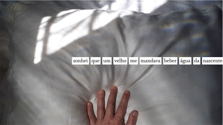Foto horizontal colorida mostrando uma mão sobre um travesseiro branco. A mão está centralizada e cortada na margem de baixo, deixando ver apenas os dedos. O travesseiro ocupa quase todo o quadro. Há um retângulo enviezado de luz do sol atravessando a parte de cima do travesseiro. Embaixo dele e acima da mão há uma frase numa faixa branca: sonhei que um velho me mandava beber água da nascente.