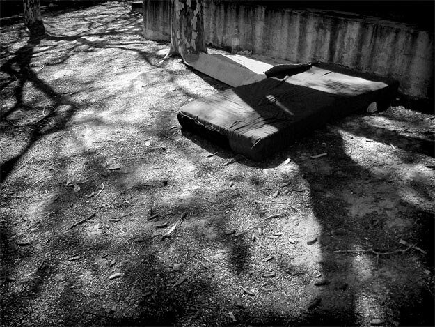 Um colchão com forro meio rasgado, estendido sobre o chão de terra, está encostado a uma parede velha e manchada, na parte de cima da foto, à direita. Ao lado dele aparece parte de um tronco de árvore. Na extrema esquerda, no alto, outro tronco de árvore. As duas árvores lançam sombras de galhos no chão, coberto folhinhas caídas, gravetos e sementes.