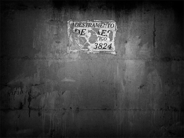 Foto de um cartaz quase destruído, branco com letras pretas, pequeno, na parte de cima da imagem, ao centro. A parede é escura, com manchas mais claras e outras mais escuras, de algo que parece ter escorrido. Na parte de cima e nas margens laterais, a imagem está quase preta; a pouca luz está no cartaz e na parte central. O pequeno cartaz foi colado e depois teve várias partes arrancadas; com alguma dificudade, pode-se concluir que dizia "adestramento de cães", junto a um número de telefone.(Esta é a última imagem da série.) 