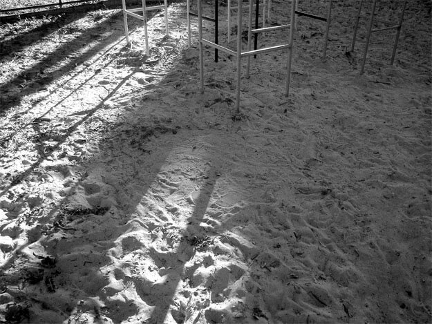 O chão de areia branca de parque infantil ocupa todo o quadro, com uma faixa de luz do sol na metade da esquerda. No alto, à direita, vê-se a parte de baixo de um trepa-trepa, que lança sombras diagonais sobre a parte iluminada.