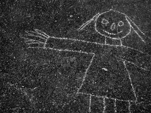 Detalhe de uma boneca ou menina desenhada com giz branco sobre uma superfície de asfalto escura, quase preta, com pedrinhas brancas muito pequenas. O desenho parece ter sido feito por uma criança.