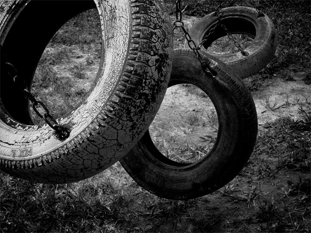 Detalhe de três balanços de pneus pendurados por correntes, enquadrados da esquerda para a direita e de cima para baixo. Eles ocupam todo o quadro, exceto pelo chão, que era de areia mas está tomado por capim baixo. Nenhum dos pneus aparece por inteiro, pois o maior está cortado acima e à esquerda, o segundo está parcialmente encoberto pelo primeiro e o terceiro está parcialmente encoberto pelo segundo. Eles parecem ter recebido pintura em tons diferentes, que está desgastada pelo tempo.