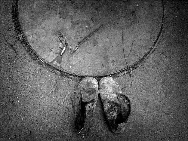 Detalhe da tampa de um bueiro e de duas botas velhas sobre um chão de cimento. O bueiro forma um semicírculo na parte de cima da foto, e está tampado. Na parte de baixo, ao centro, há duas botas de couro escuro. A ponta das botas toca a tampa do bueiro. Elas estão muito usadas e arranhadas, sem cadarços e manchadas com um pó branco.
