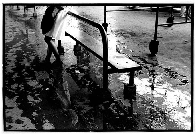 Foto horizontal de uma mulher de pé, de perfil, encostada a um banco de metal. Ela aparece só da cintura para baixo; usa saia e blusa brancas, uma grande bolsa escura e sapatilhas escuras. Grande parte do piso está coberta por poças d'água compridas. Abaixo da mulher vê-se parte de seu reflexo na água. O banco é longo e há outro igual ao longe, do qual se vê apenas as pernas. Há também as pernas de um andaime sobre grandes rodízios, em frente ao banco. A foto é escura, exceto pela saia da mulher, o assento do banco e algumas poças d'água no chão; a luz que bate nesses elementos alinha-os numa faixa diagonal clara.