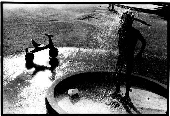 Foto horizontal de uma criança de cerca de cinco anos. Ela está embaixo de um filete de água que cai sobre sua cabeça e se espalha para os lados. Só a sua silhueta é visível, preta na contraluz. Ele está do lado direito da foto, virado para a câmera em meio perfil. A água cai sobre uma área redonda delimitada por uma borda de concreto, cortada embaixo e à direita. A foto é escura; o chão é de asfalto e há uma parte molhada no lado esquerdo, refletindo a luz que vem de cima. Sobre essa parte molhada, branca, há um velotrol em silheta e seu reflexo na água.