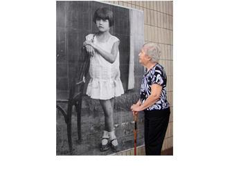 Foto vertical de uma idosa olhando o retrato antigo de uma menina, colado na parede. A idosa está de perfil, segura uma bengala de madeira com as duas mãos juntas em frente ao corpo, veste uma calça preta e uma blusa preta e branca. Ela está olhando para cima, para o rosto da menina. Na foto antiga a menina aparece em tamanho um pouco maior que o natural. Ela está de pé num chão de terra batida, junto a uma cadeira de madeira. Tem cabelos curtos e lisos, com corte chanel. Usa um vestido branco curto, de cintura baixa, sapatos boneca bem usados e meias brancas curtas. Suas pernas estão sujas. Ela apoia um dos braços no encosto da cadeira e suas mãos estão juntas. Está ligeiramente de perfil, com o olhar ao longe, e tem as pernas cruzadas, numa pose harmônica e claramente arranjada pelo fotógrafo. No cantinho da foto está escrito "1929", e, embaixo, "Estava c/ 6 anos".