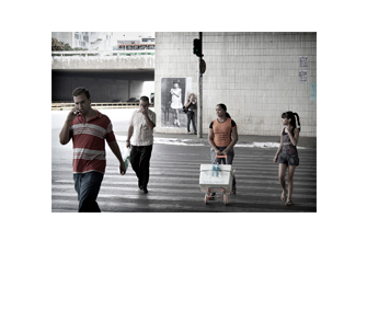 Foto horizontal do mesmo local da imagem anterior, agora enquadrado de frente e de longe, do outro lado da rua. Quatro pessoas atravessam a faixa de segurança, de frente para a câmera: um homem jovem falando ao celular; um homem de cabeça baixa, apertando os olhos com a mão; uma vendedora de água, empurrando um isopor num carrinho; e uma menina de 10 ou 11 anos. Do outro lado da rua vê-se o paredão onde está colada a foto antiga, no canto esquerdo da parede e no centro da foto maior. Encostada à foto antiga há uma mulher jovem, em meio perfil, e à sua frente um poste com um semáforo. Ao fundo vê-se um viaduto, ônibus, carros e prédios.