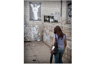 Foto vertical de uma mulher passando de costas para a câmera, à direita. Ela tem cabelos lisos e compridos, avermelhados; usa jeans e blusa lilás, carrega uma grande bolsa e um guarda-chuva. Está andando devagar, cabisbaixa, numa calçada de cimento. Do seu lado direito há rampa; à frente, uma parede alta que ocupa todo o fundo da foto. No canto esquerdo da parede, em cima, está colada uma grande foto vertical de uma menina em pé, de mãos postas, olhando para a câmera, com um vestido branco e longo de mangas compridas, cabelo louro comprido, asas e auréola de anjo. A parede do fundo e a da rampa são revestidas de ladrilhos antigos e muito sujos. Há vários cartazes colados, alguns sobre outros, em diferentes estados, alguns já totalmente arrancados. Nenhum foi colado em cima da foto antiga; eles respeitam uma certa distância e não passam da metade da sua altura, pois a foto está bem alta. 