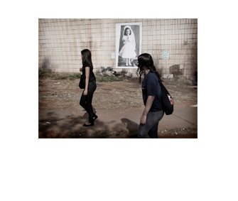 Foto horizontal de duas jovens passando de perfil, com os rostos ocultos, da direita para a esquerda. Elas têm cabelo comprido e escuro e usam roupas escuras; estão afastadas uma da outra. Vê-se uma calçada de cimento e depois o chão de terra com mato seco aparado. Da metade da foto para cima, enquadrado de frente, há um paredão revestido de ladrilhos claros, velhos e sujos. A parede tem grandes manchas escuras.Entre as duas moças vê-se uma foto vertical colada na parede, a cerca de oito ou nove metros de distância da câmera, mostrando uma menina de cabelos escuros, com seis ou sete anos, sorrindo e olhando ligeiramente para o lado. Está de mãos postas e toda de branço: vestido, véu e grinalda, luvas, meias e sapatos.