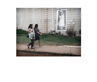 Foto horizontal de duas mulheres passando juntas na calçada, da esquerda para a direita, de perfil, olhando para a foto da menina. A foto antiga está enquadrada em perspectiva e mais de perto que na imagem anterior.