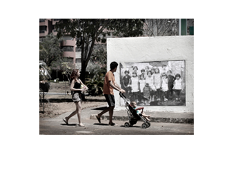 Foto horizontal de um casal passando com um bebê num carrinho, todos de perfil, olhando uma foto antiga colada na parede. A foto colada tem cerca de 3m x 2m e mostra um grupo de 14 crianças, que têm entre 4 e 11 anos de idade. As crianças posam olhando para a câmera. Todas estão de pé, menos a mais velha; ela está sentada no centro do grupo e segura pela mão uma bebê de dois anos ou menos, que está em pé, de vestido e touca. Na foto maior, atrás da parede, aparece um gramado, uma calçada, uma árvore, algumas plantas e um prédio de apartamentos ao fundo.