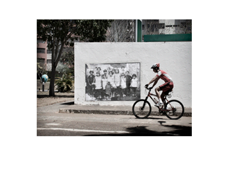 Foto horizontal de um homem passando de bicicleta, de lado para a câmera, da direita para a esquerda. Ele usa roupas de ciclista e um capacete de formato futurista, próprio para esse esporte. Suas roupas, capacete e bicicleta são vermelhas com detalhes brancos. Está em frente à parede onde está colada uma foto antiga (a mesma das imagens anteriores). A roda da frente da bicicleta está junto ao canto da foto antiga. Atrás da parede vê-se uma árvore, um orelhão, algumas plantas e dois prédios.