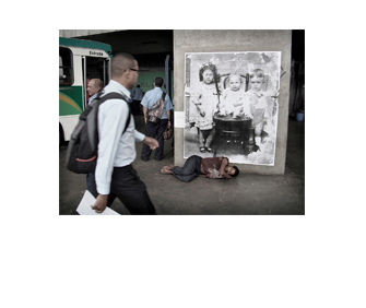 Foto horizontal do mesmo local, com o mesmo enquadramento. Junto à coluna com o retrato das crianças há um homem dormindo no chão, de lado, descalço. O lado de cima do seu corpo está sobre a margem inferior da foto antiga. Bem perto da câmera há um homem passando no lado esquerdo, indo para a direita e olhando a foto. Atrás dele vê-se parte de um ônibus, dois homens com camisa de uniforme azul claro e pessoas no fundo, ao longe.