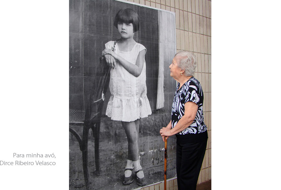 Foto vertical de uma idosa olhando o retrato antigo de uma menina, colado na parede. A idosa está de perfil, segura uma bengala de madeira com as duas mãos juntas em frente ao corpo, veste uma calça preta e uma blusa preta e branca. Ela está olhando para cima, para o rosto da menina. Na foto antiga a menina aparece em tamanho um pouco maior que o natural. Ela está de pé num chão de terra batida, junto a uma cadeira de madeira. Tem cabelos curtos e lisos, com corte chanel. Usa um vestido branco curto, de cintura baixa, sapatos boneca bem usados e meias brancas curtas. Suas pernas estão sujas. Ela apoia um dos braços no encosto da cadeira e suas mãos estão juntas. Está ligeiramente de perfil, com o olhar ao longe, e tem as pernas cruzadas, numa pose harmônica e claramente arranjada pelo fotógrafo. No cantinho da foto está escrito "1929", e, embaixo, "Estava c/ 6 anos".